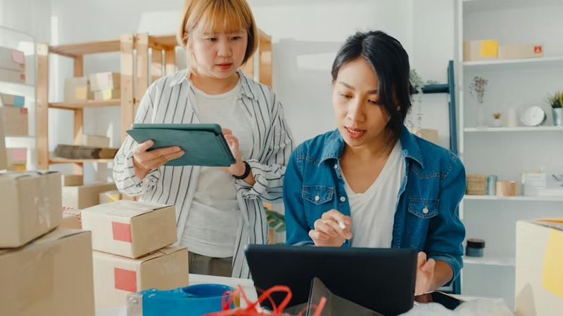 Two women in a room filled with boxes, working with their gadgets.