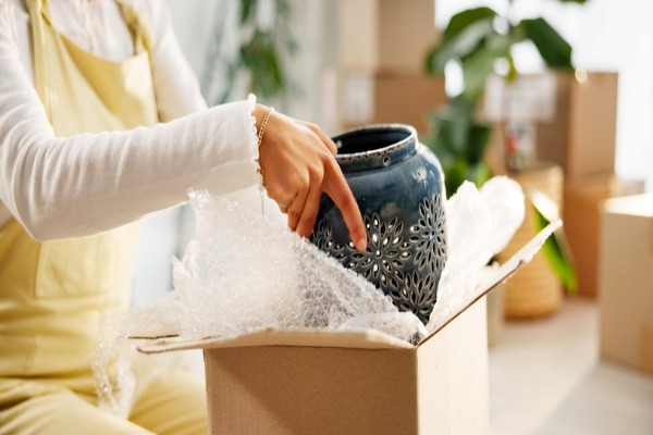 Person carefully placing a decorative ceramic vase into a cardboard box with bubble wrap for protective packaging during shipping or moving.