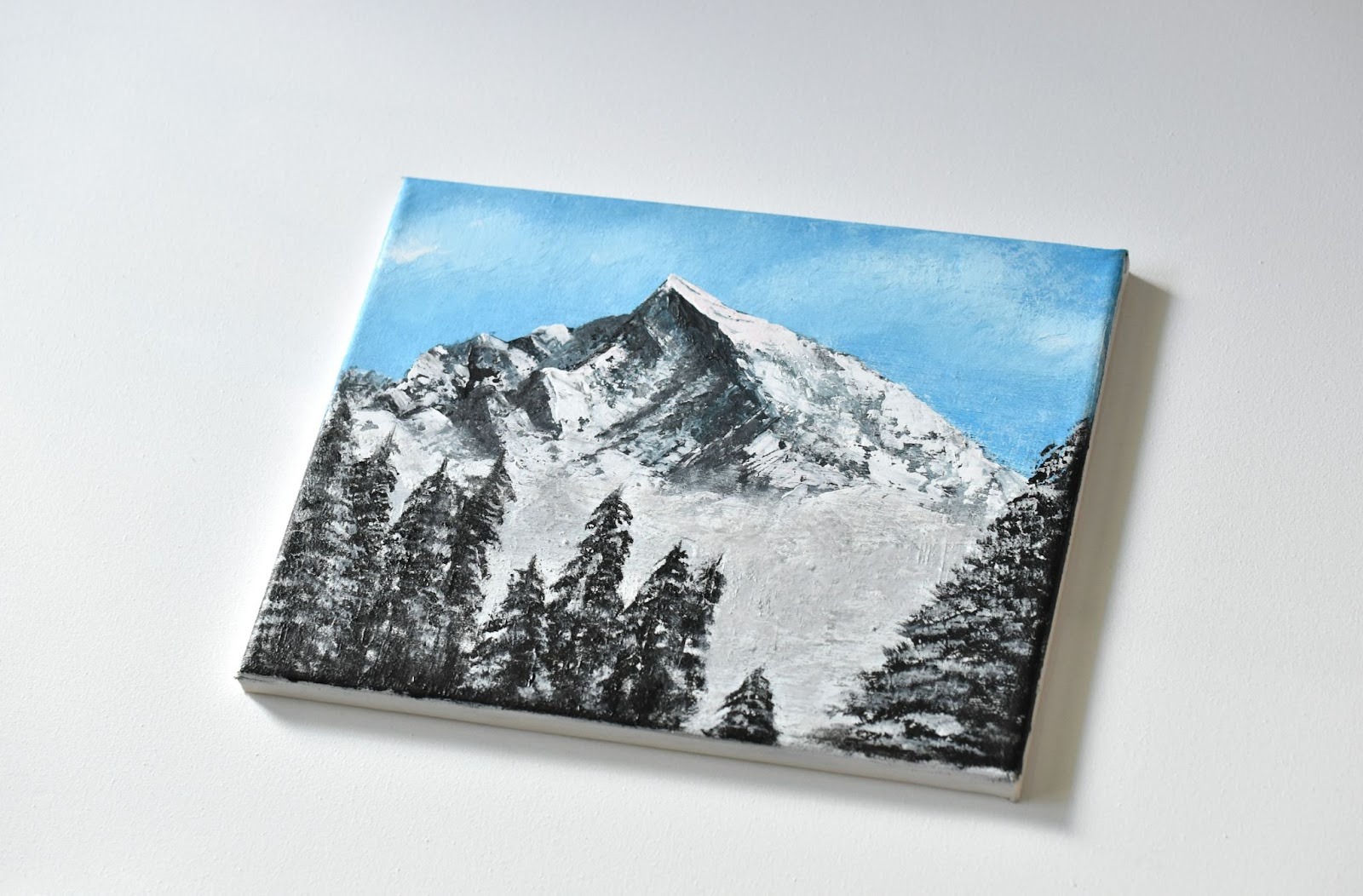 Scenic painting of a mountain with trees at the bottom, painted over a canvas