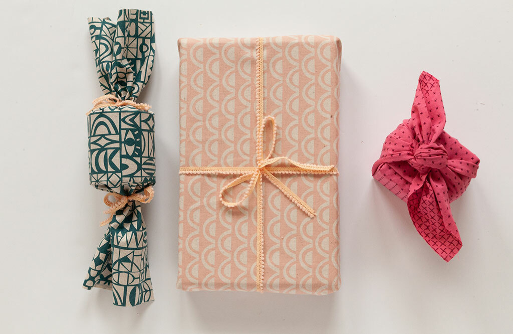 Three gifts wrapped furoshiki-style in teal, peach, and pink patterned fabric squares with ribbon ties.