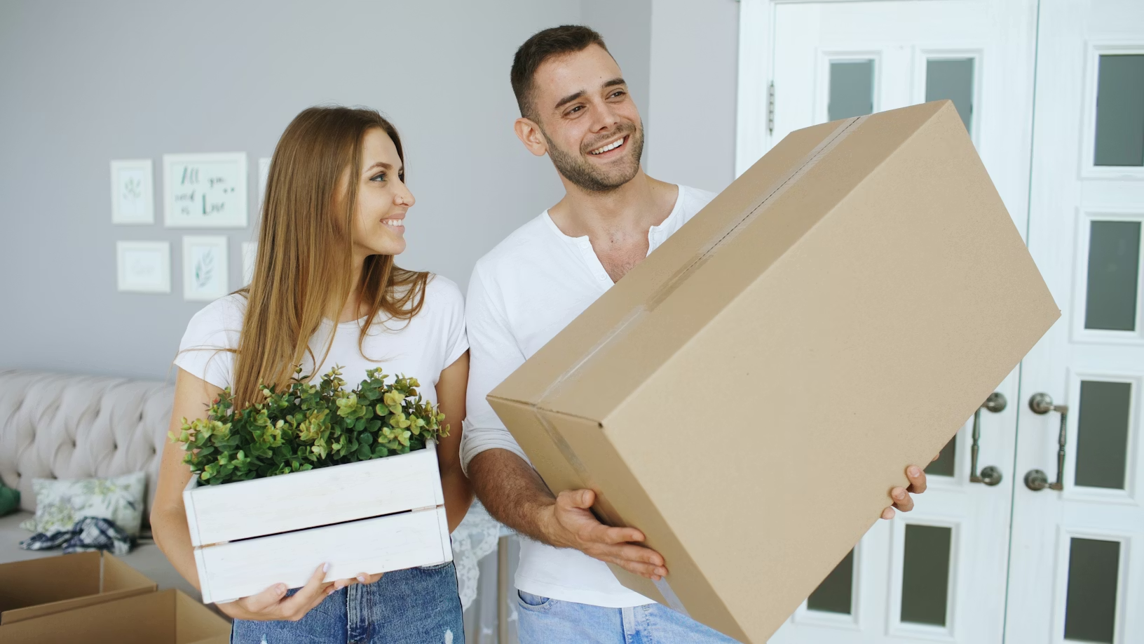  Young happy couple smiling while holding an artificial flower box and a tightly sealed cardboard box. 