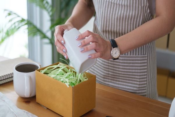 Close-up of hands placing a white ceramic bottle into a box with shredded filler.