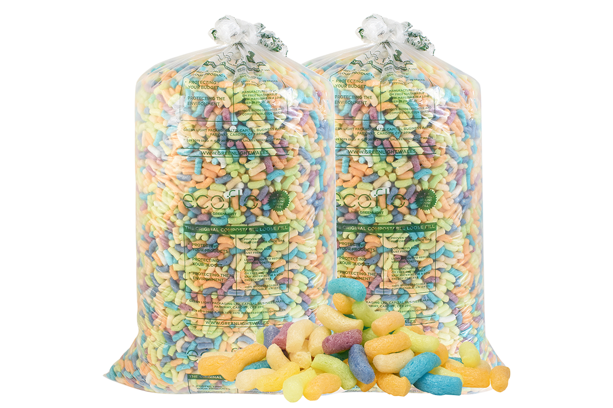 Two large clear plastic bags filled with multicolored biodegradable packing peanuts in pink, blue, green, yellow, and orange, with a small pile of loose packing peanuts spilled in front.