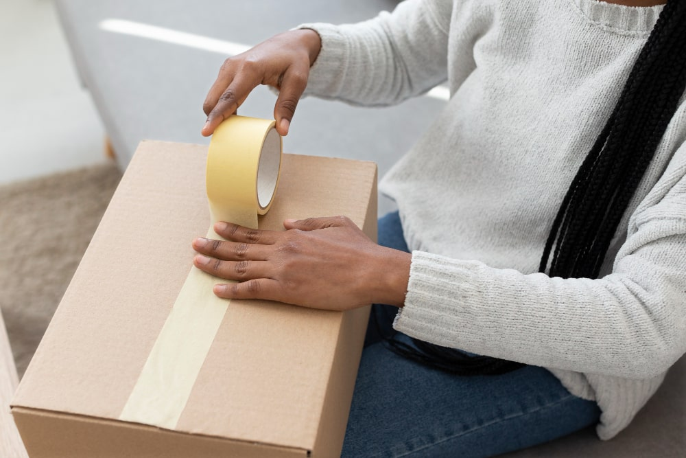 Woman sealing a cardboard shipping box with a roll of kraft paper tape, showing it in practical use.