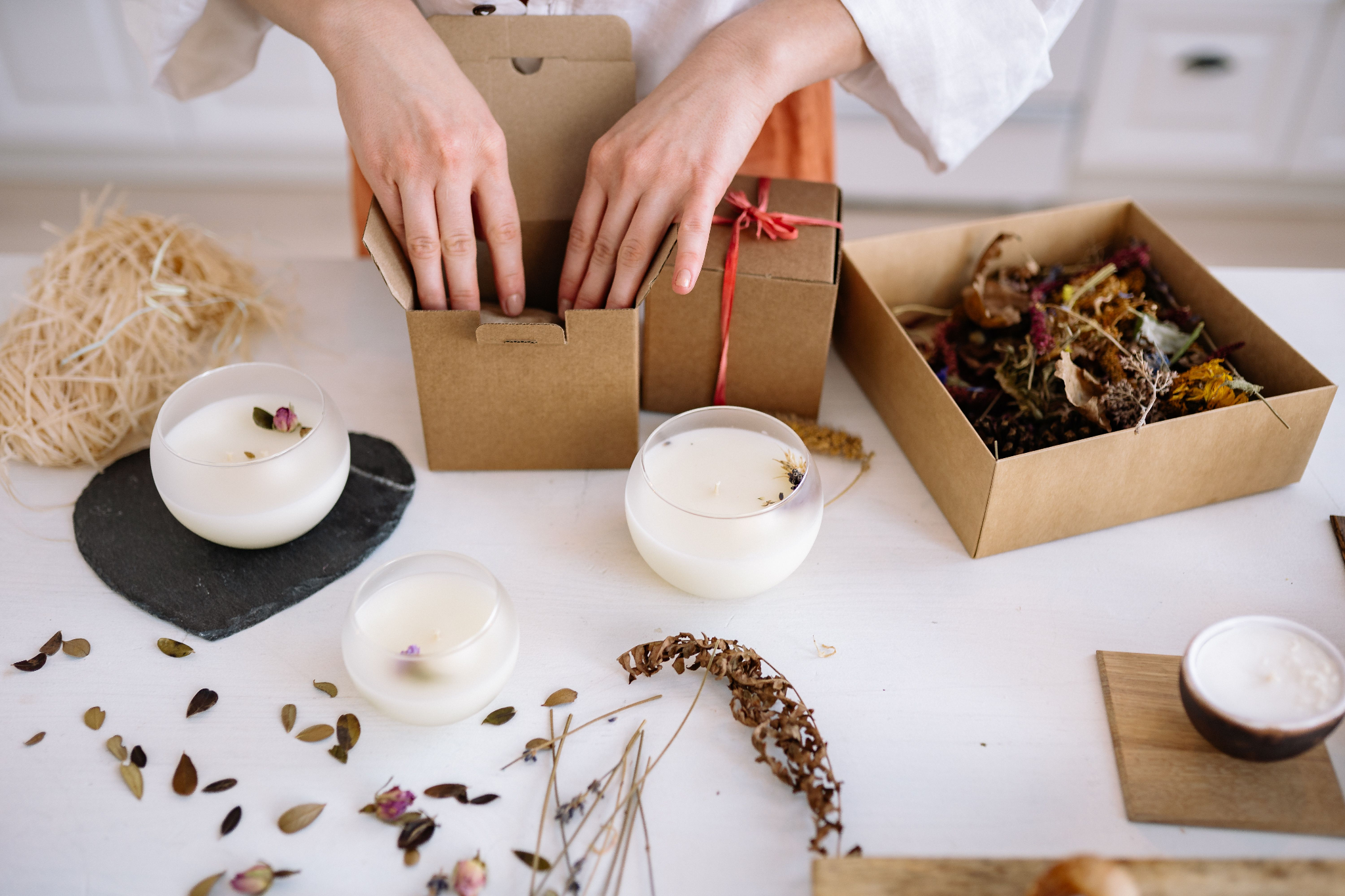 Hands packing handmade candles into kraft gift boxes alongside dried flowers and botanicals on a white table.