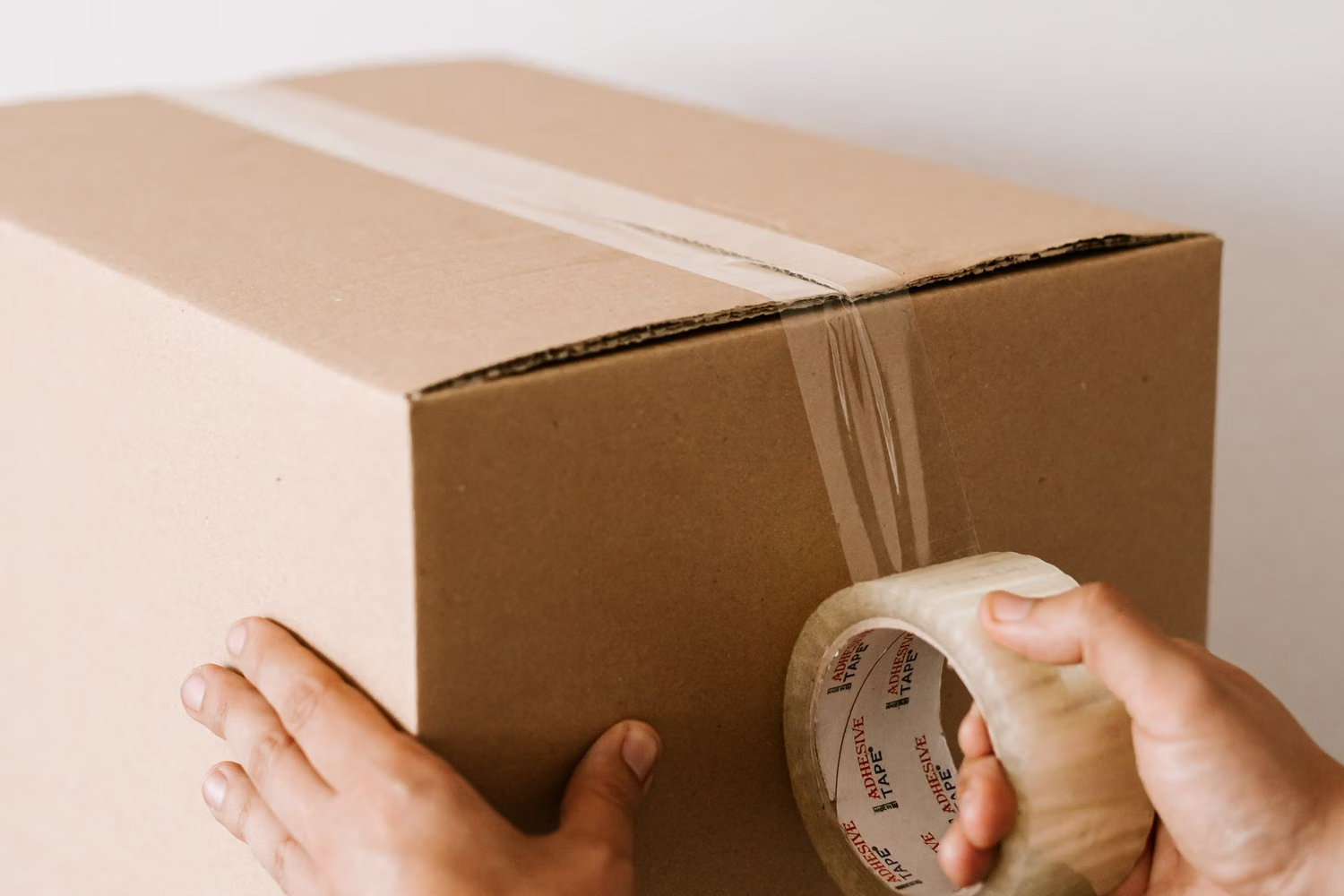 Hands sealing a cardboard box with a roll of clear adhesive packing tape.