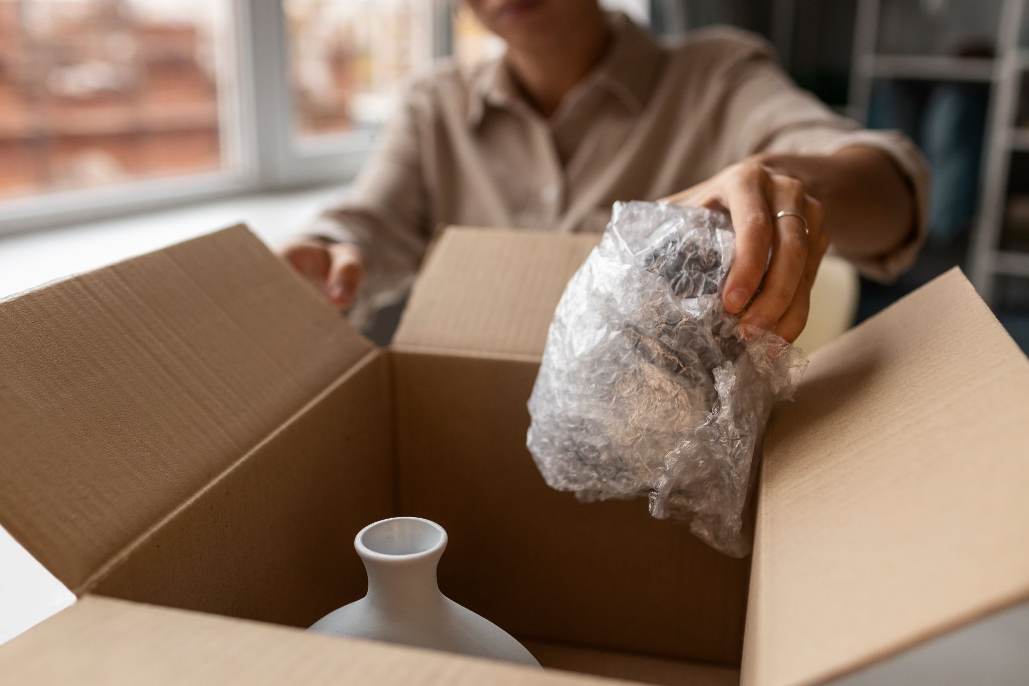 Person placing a bubble-wrapped item into a cardboard box beside a white ceramic vase.