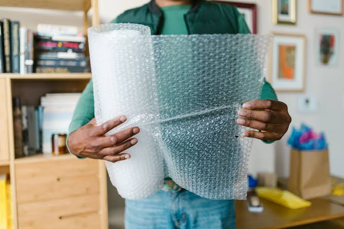 Person in green shirt holding a roll of clear bubble wrap indoors near a bookshelf and packing area.