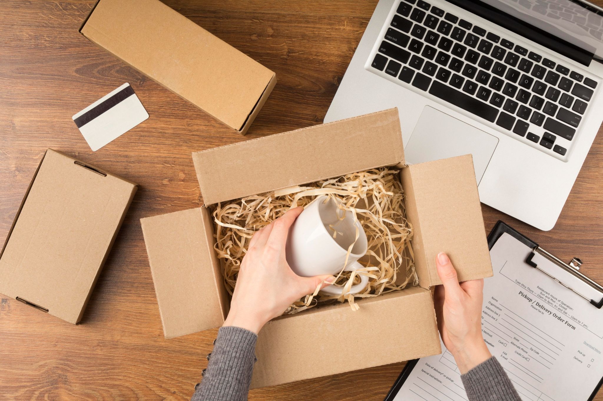 Overhead view of hands placing a white mug into a cardboard box filled with shredded paper void fill.
